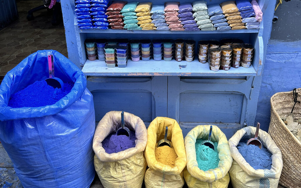 Bright, colorful open sacks of powdered indigo blue, yellow, and red paint pigments displayed on a stone street in the Chefchaouen medina, Morocco.