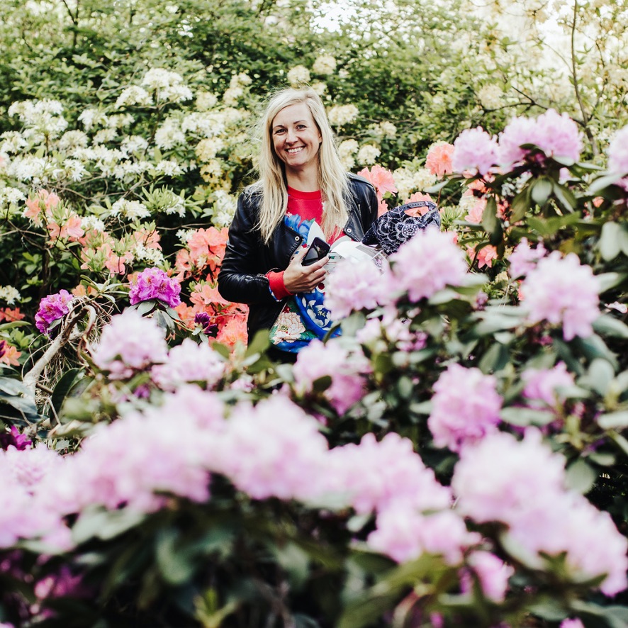 The author of Morocco Notes, a blonde woman in a black leather jacket, smiling among vibrant rhododendron flowers.