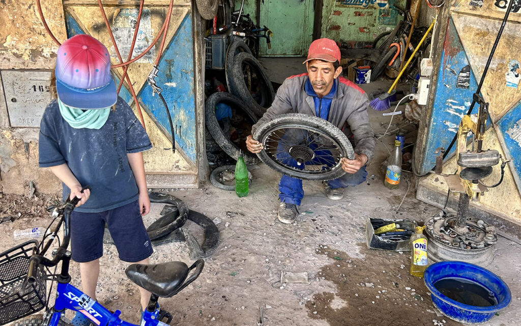 A local Moroccan mechanic repairing a bicycle wheel for a young traveler in a small workshop.