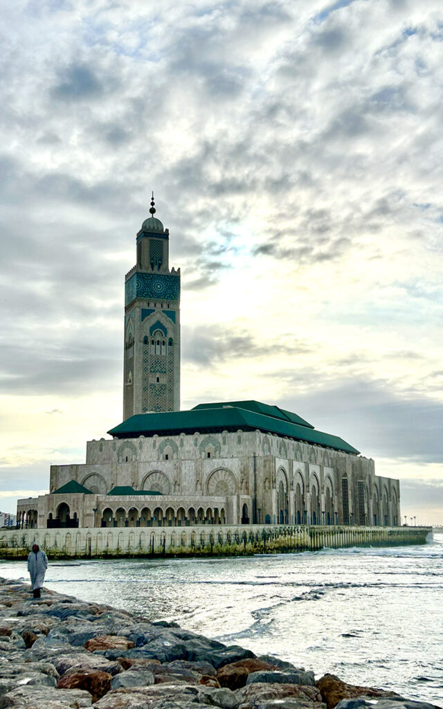 Hassan II Mosque in Casablanca from the ocean side with waves and a person walking along the rocks