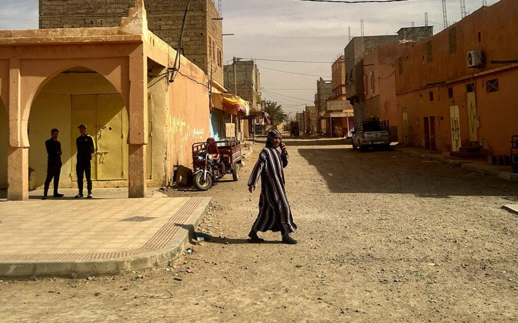 A Moroccan man wearing a traditional striped djellaba walking across a dusty street in a small town.