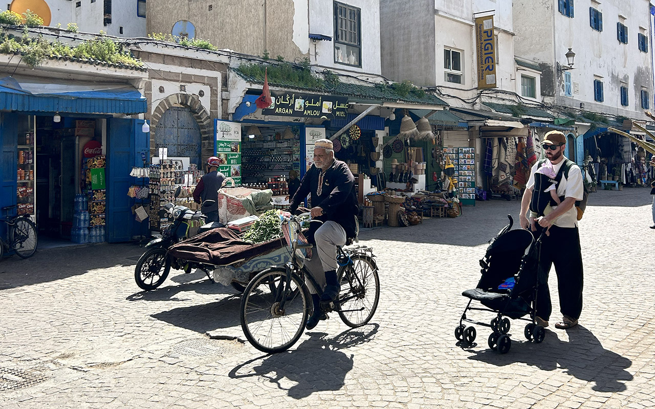 A busy Moroccan street market with a local man on a bicycle and a tourist pushing a stroller.