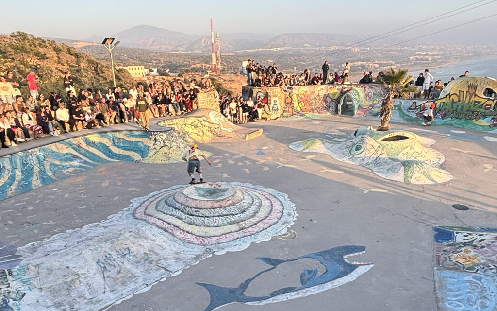 Wide view of Taghazout skatepark with colorful graffiti murals and crowd watching a skater, Morocco