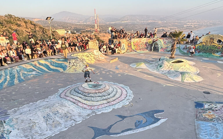 Wide view of Taghazout skatepark with colorful graffiti murals and crowd watching a skater, Morocco