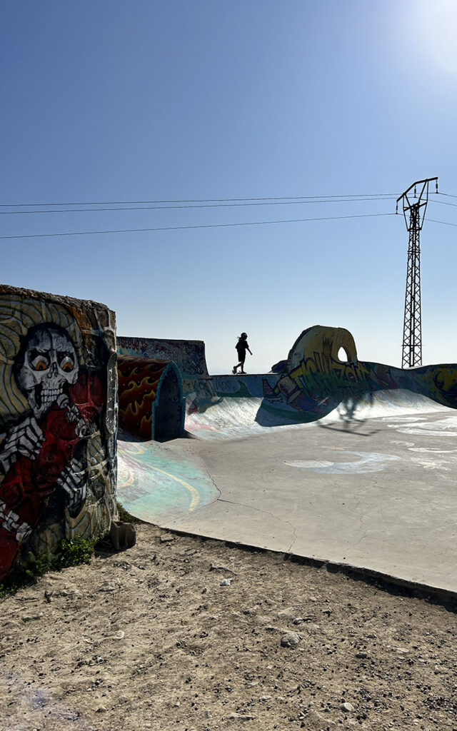Skater silhouette against blue sky at Taghazout skatepark with skull graffiti mural, Morocco