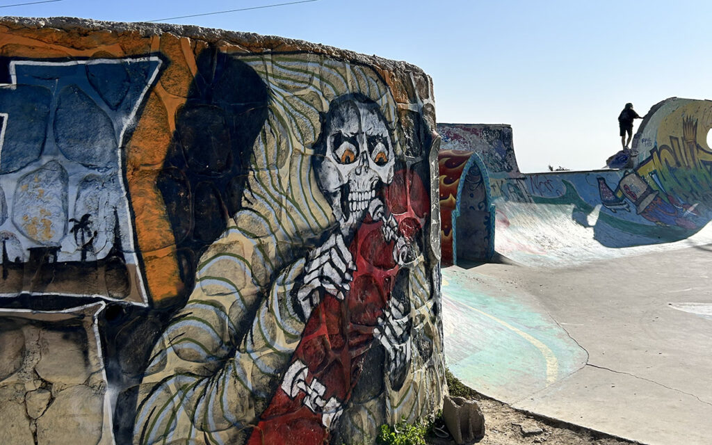 Skater silhouette against blue sky at Taghazout skatepark with skull graffiti mural, Morocco
