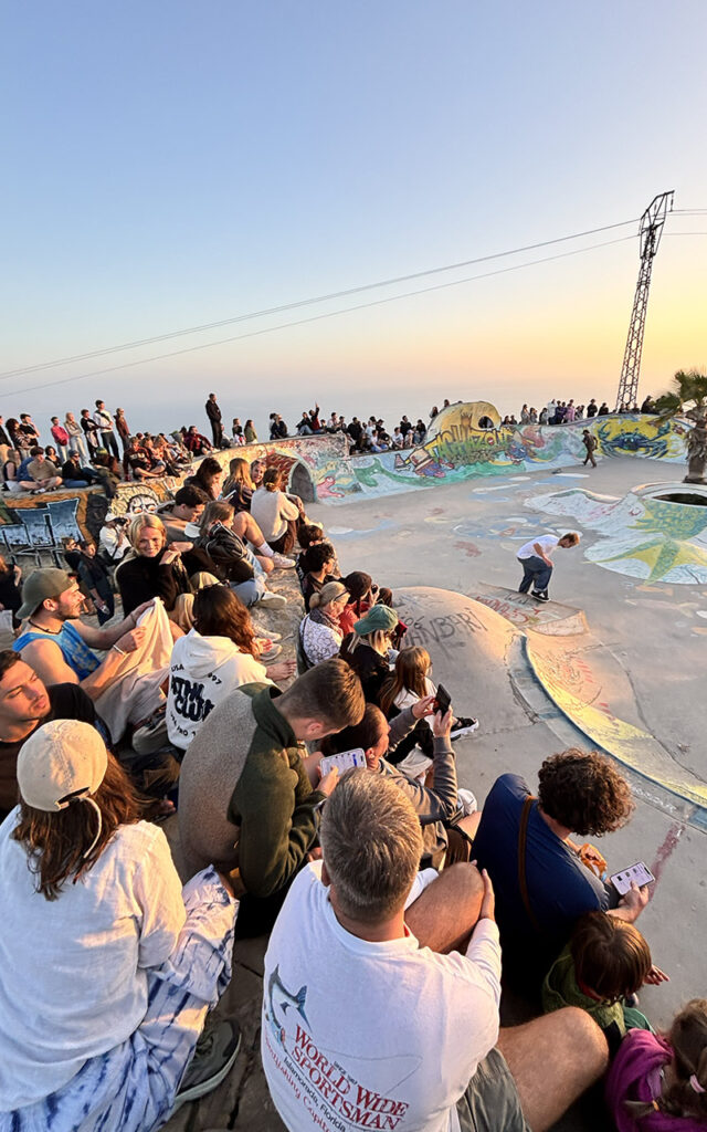 People sitting and watching skating at Taghazout skatepark hillside at dusk, Morocco