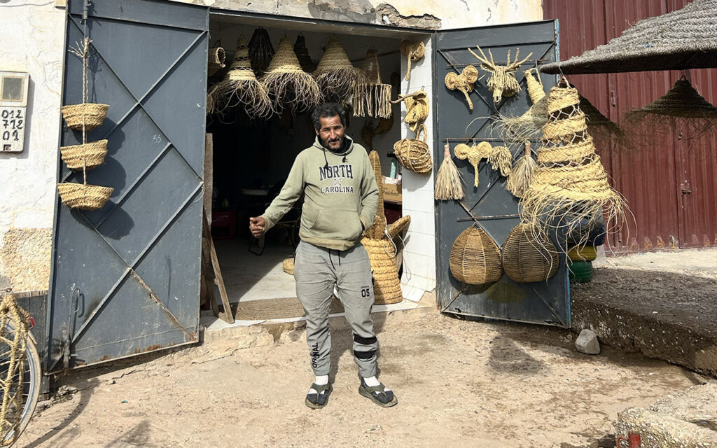 A Moroccan craftsman standing in front of his workshop filled with traditional handmade wicker baskets and lamps.