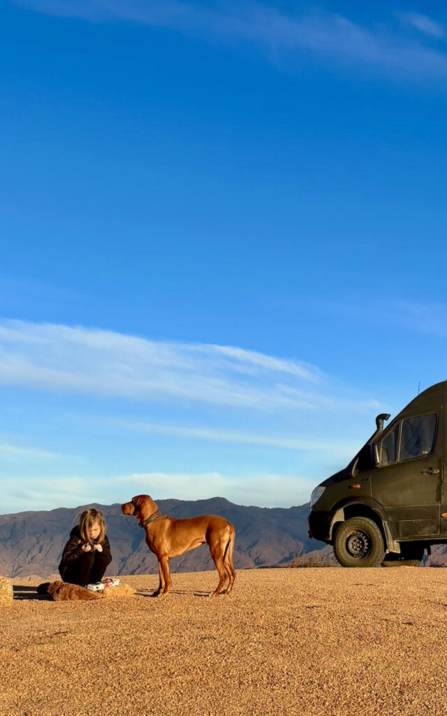 A child and a Vizsla dog standing by an off-road camper van with the Atlas Mountains in the background at sunset