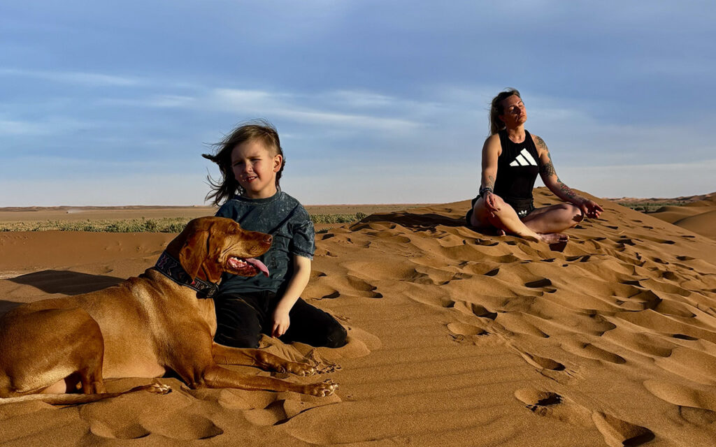 Hungarian Vizsla dog Flēra and child on sand dunes in Sahara desert, with woman meditating in the background - family vanlife in Morocco