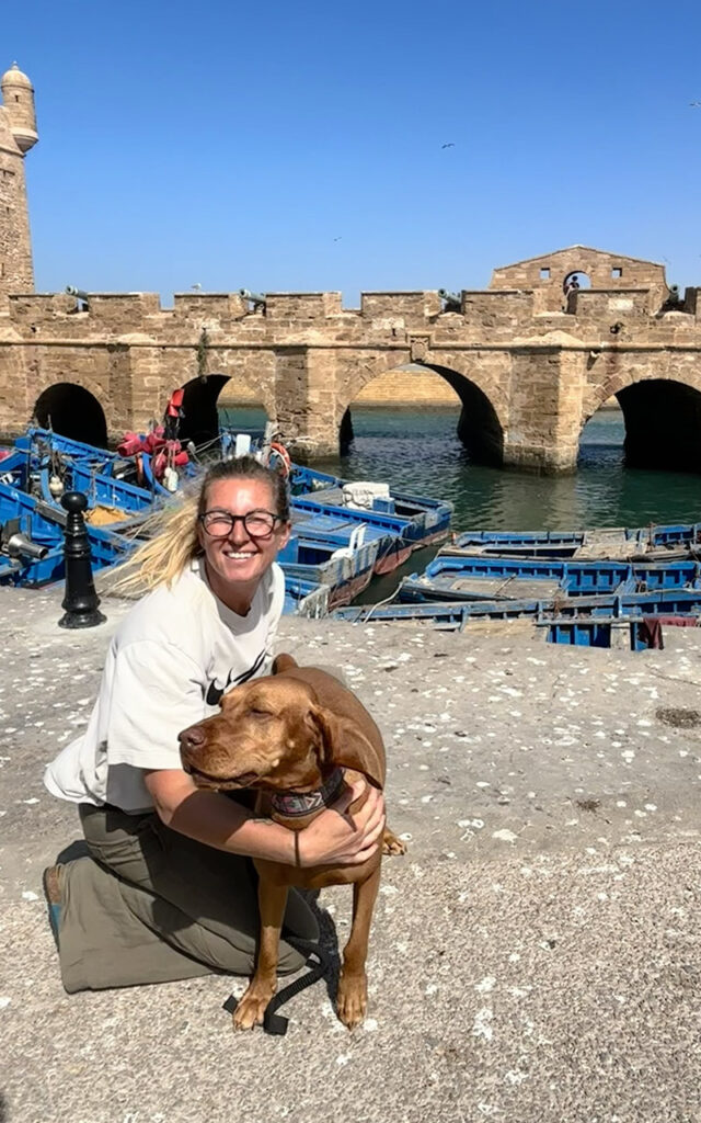 A woman hugging her dog in front of the historic sea bastions and blue fishing boats in Essaouira, Morocco